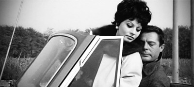 A black and white photo of Sophia Loren and Marcello Mastroianni in a Rolls-Royce car while they swap their place in the seats. The scene is from the second episode of Yesterday, Today and Tomorrow